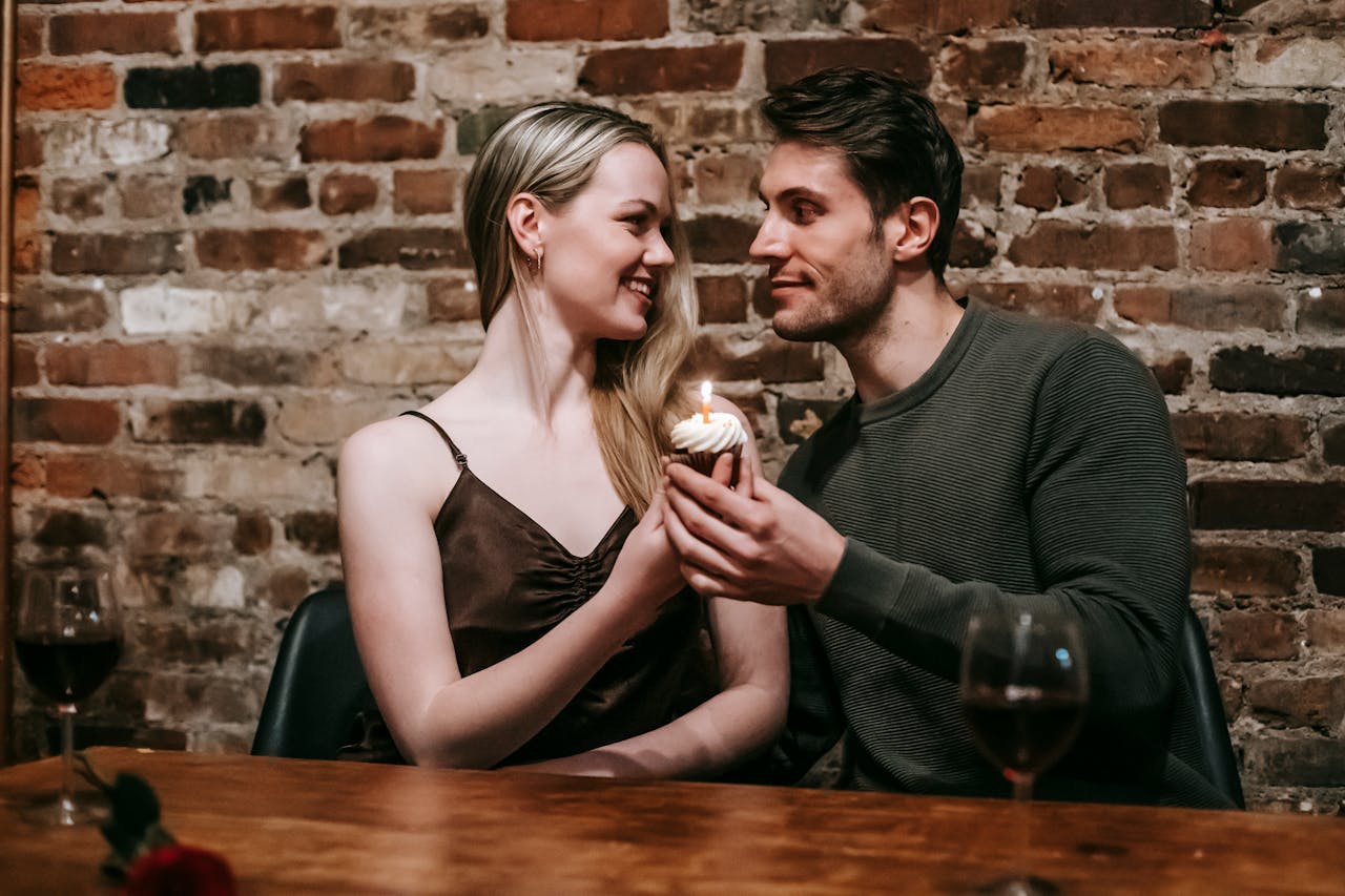 Happy young couple in elegant clothes sitting at table and celebrating birthday with small cupcake and candle near glasses with red wine and brick wall while looking at each other